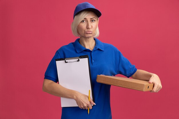 Triste livreuse blonde d'âge moyen en uniforme bleu et casquette tenant un paquet de pizza au crayon presse-papiers isolé sur un mur rose avec espace de copie