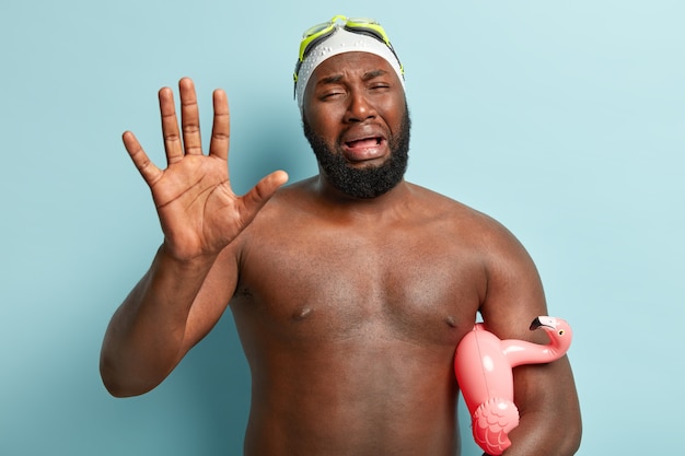 Triste mécontent homme à la peau sombre pleure de panique, montre la paume, frustré d'avoir gâté les vacances d'été, porte des lunettes, tient l'anneau de natation flamingo, isolé sur mur bleu