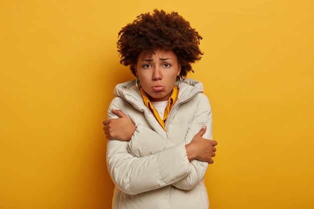 Triste mécontentement femme frisée se sent gelée, tremble de froid, porte des vêtements d'extérieur blancs, croise les bras sur la poitrine, se dresse sur fond de studio jaune