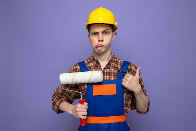 Triste montrant le pouce vers le haut d'un jeune constructeur masculin portant un uniforme tenant une brosse à rouleau