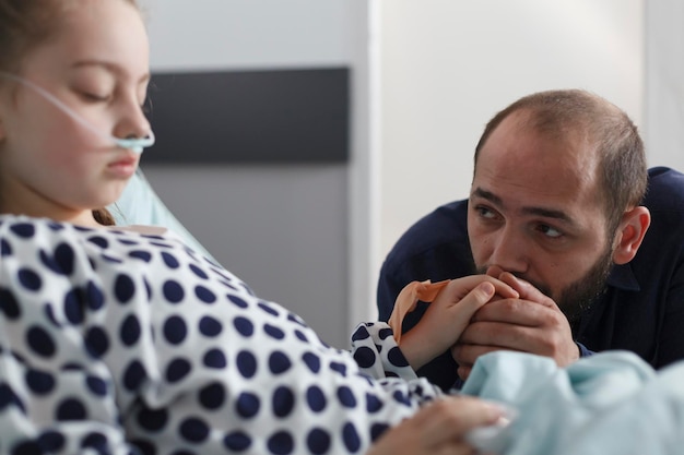 Triste père attentionné anxieux tenant la main de petits enfants malades endormis dans le service des patients de l'hôpital pédiatrique. Fille malade utilisant un tube nasal dans un lit d'hôpital pendant qu'un parent attentif la regarde.