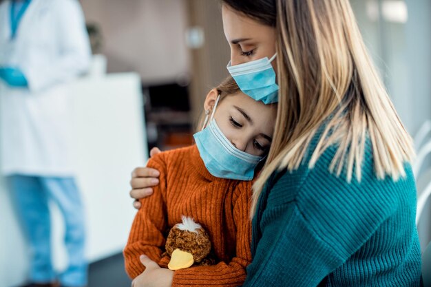 Triste petite fille assise sur les genoux de sa mère alors qu'elle était assise dans la salle d'attente d'une clinique médicale pendant la pandémie de coronavirus