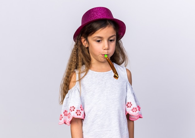 triste petite fille caucasienne avec un chapeau de fête violet soufflant un sifflet de fête isolé sur un mur blanc avec un espace de copie