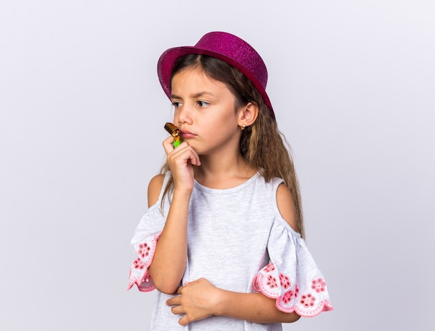 Triste petite fille de race blanche avec chapeau de fête pourpre tenant un sifflet de fête et regardant à côté isolé sur un mur blanc avec espace de copie