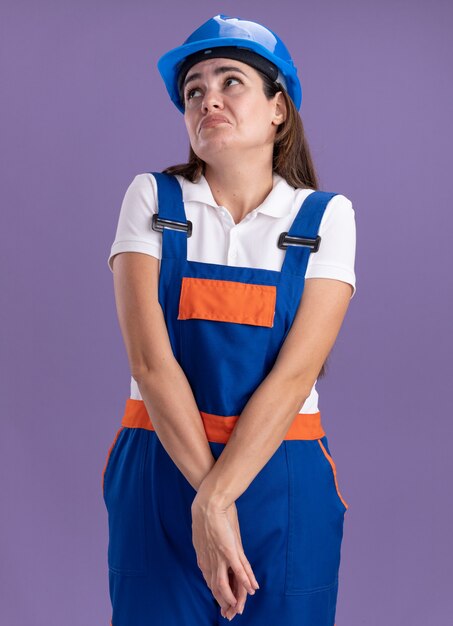 Triste En Regardant La Jeune Femme De Constructeur En Uniforme Tenant Les Mains Ensemble Isolées Sur Le Mur Violet