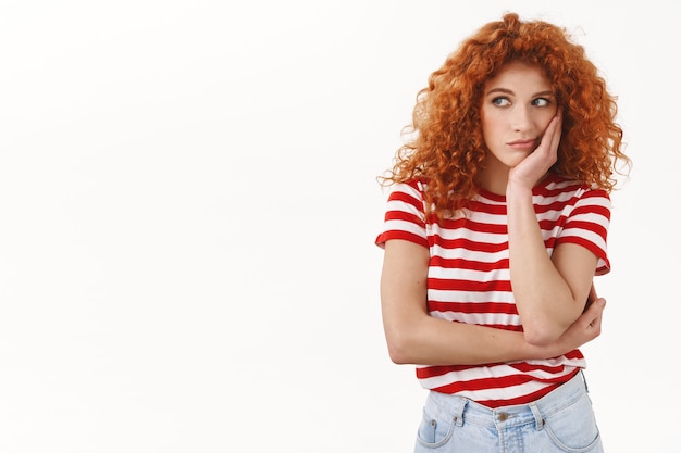 Triste rousse européenne femme frisée tête maigre paume ennui regret regarder à gauche jaloux assis bureau ennuyeux fête de rêve debout bouleversé malheureux t-shirt rayé mur blanc réticent