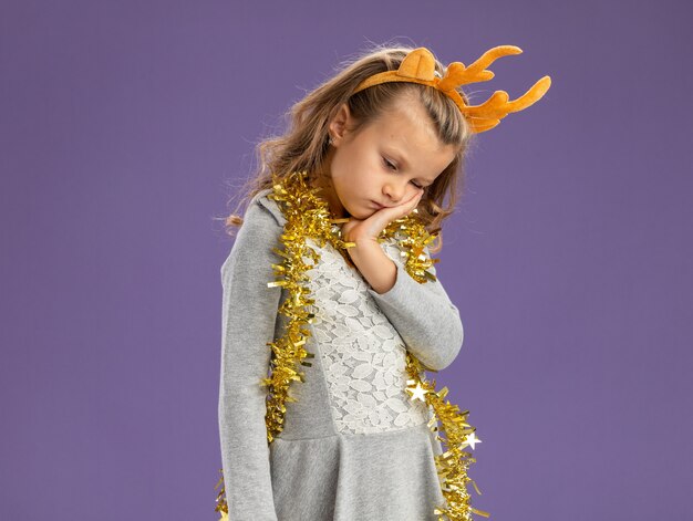 Triste avec tête baissée petite fille portant un cerceau de cheveux de Noël avec guirlande sur le cou mettant la main sur la joue isolé sur fond bleu avec copie espace