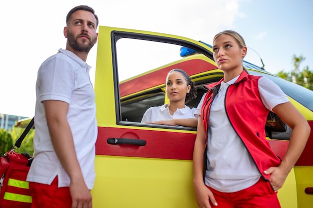 Trois ambulanciers debout près de l'ambulance parlant Paramédic masculin portant un sac de traumatologie médicale Groupe de trois ambulanciers debout devant l'ambulance avec le sourire