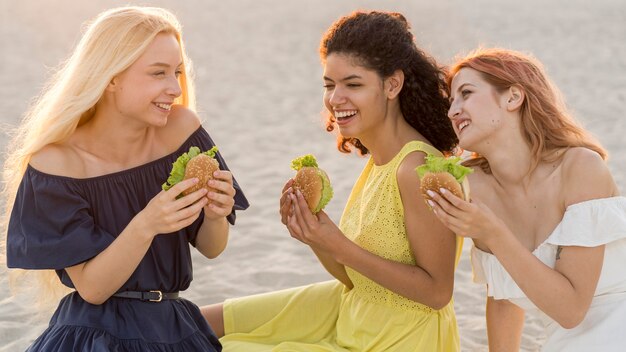 Trois amies appréciant des hamburgers à la plage ensemble