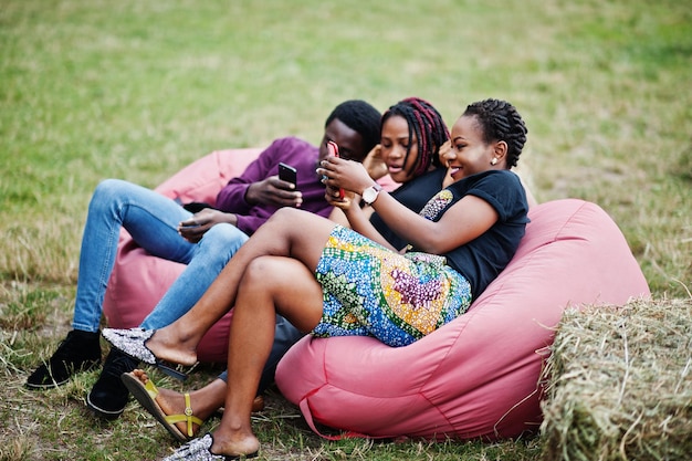 Photo gratuite trois amis afro-américains se détendent assis sur des poufs et utilisent leur téléphone à l'extérieur