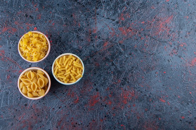 Trois assiettes colorées avec différents types de pâtes non cuites sur un fond sombre.