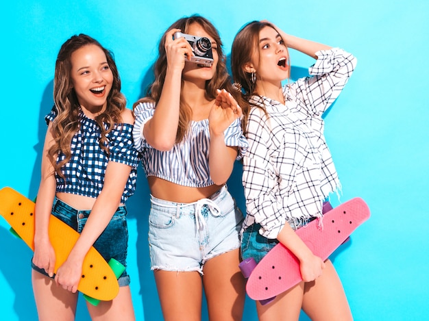 Trois belles filles souriantes élégantes et sexy avec des planches à roulettes colorées Femmes en vêtements de chemise à carreaux d'été posant. Modèles prenant des photos sur un appareil photo rétro