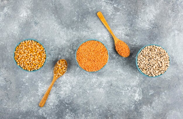 Trois bols de grains de maïs, de haricots et de lentilles rouges sur une surface en marbre.