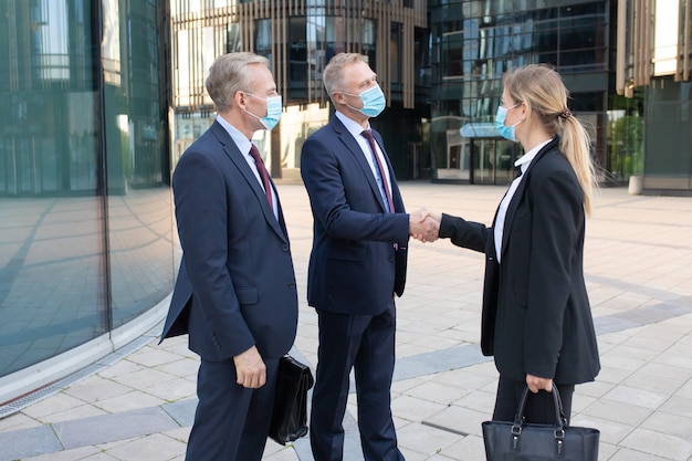 Trois employés de bureau dans des masques faciaux faisant affaire ou salutation. Femme d'affaires et hommes d'affaires prospères professionnels debout à l'extérieur et poignée de main. Concept de négociation, de protection et de partenariat