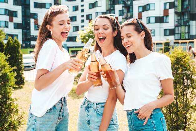Trois jeunes belles femmes hipster souriantes dans les mêmes vêtements d'été à la modeFemmes insouciantes sexy posant dans la rueModèles positifs s'amusant dans des lunettes de soleil Boire une bouteille de bière Oktoberfest