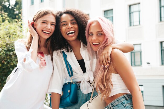 Trois jeunes belles femmes souriantes dans des vêtements d'été à la modeFemmes multiraciales insouciantes sexy posant sur le fond de la rueModèles positifs s'amusant dans des lunettes de soleil Enthousiaste et heureux