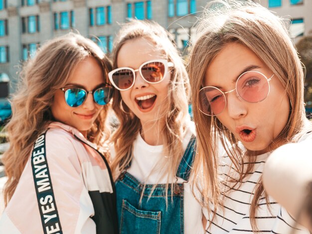 Trois jeunes femmes souriantes hipster dans des vêtements d'été.Filles prenant des photos d'autoportrait selfie sur smartphone.Modèles posant dans la rue.Femme montrant des émotions positives