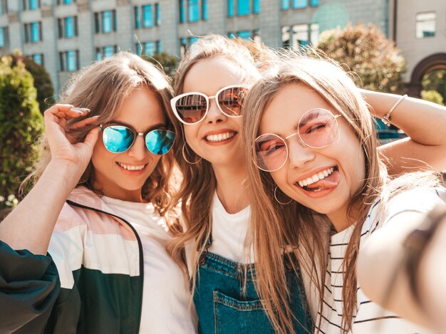 Trois jeunes femmes souriantes hipster dans des vêtements d'été.Filles prenant des photos d'autoportrait selfie sur smartphone.Modèles posant dans la rue.Femme montrant des émotions positives