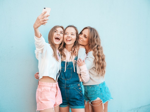 Trois jeunes femmes souriantes hipster en vêtements d'été.Filles prenant des photos d'autoportrait selfie sur smartphone.Modèles posant dans la rue près du mur.Femme montrant des émotions positives.