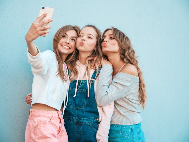 Trois jeunes femmes souriantes hipster en vêtements d'été.Filles prenant des photos d'autoportrait selfie sur smartphone.Modèles posant dans la rue près du mur.Femme montrant des émotions positives