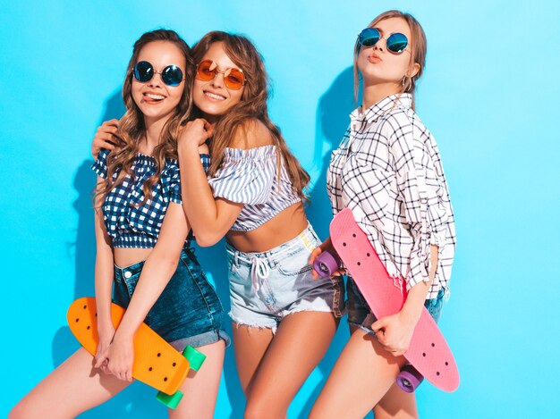 Trois jeunes filles élégantes souriantes sexy sexy avec des planches à roulettes penny colorés. Femmes en vêtements de chemise à carreaux d'été posant dans des lunettes de soleil. Modèles positifs s'amusant