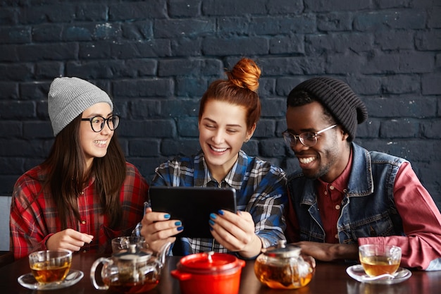 Trois jeunes gens élégants de races différentes regardant des vidéos en ligne sur une tablette numérique générique tout en dînant ensemble au restaurant