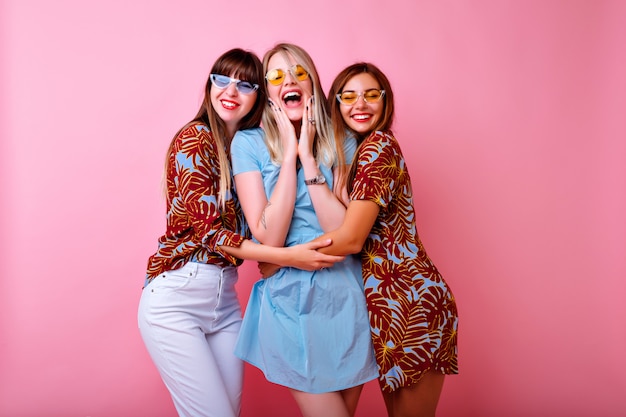 Photo gratuite trois jolies jeunes femmes heureuses faisant des selfies, un groupe de meilleurs amis s'amusant, des vêtements assortis de couleurs à la mode et des lunettes vintage, un mur rose.