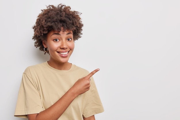 Photo gratuite tu devrais y jeter un coup d'oeil. belle femme sourit joyeusement pointe l'index dans le coin supérieur droit montre le meilleur choix promeut le produit vêtu d'un t-shirt beige décontracté isolé sur fond blanc