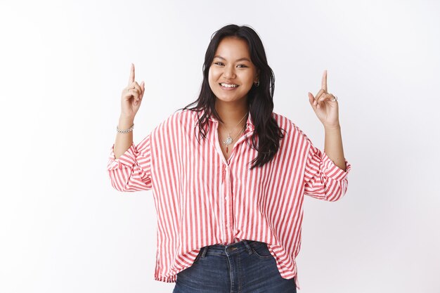 Tu ferais mieux de lever la tête. Portrait d'une jeune jolie fille asiatique enthousiaste et insouciante en blouse rayée souriante insouciante comme pointant vers le haut avec les mains levées invitant à voir une promotion intéressante