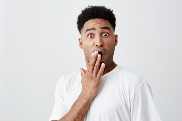 Tu ment. Émotions des peuples. Beau jeune homme américain à la peau brune avec des cheveux bouclés noirs en t-shirt blanc décontracté fermant la bouche avec la main, regardant à huis clos avec l'expression du visage choqué