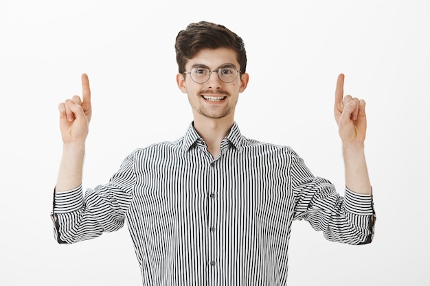 Étudiant européen amical et insouciant avec moustache et barbe dans des lunettes rondes à la mode et une chemise rayée, levant l'index et pointant vers le haut tout en souriant largement, disant qu'il a trouvé un endroit idéal