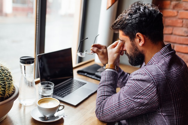 Étudiant fatigué dans des lunettes à la cafétéria, à l'aide d'un ordinateur portable