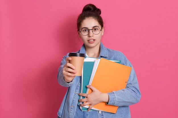 Étudiant jeune femme aux cheveux noirs offre du café à emporter, tenant un dossier papier dans les mains