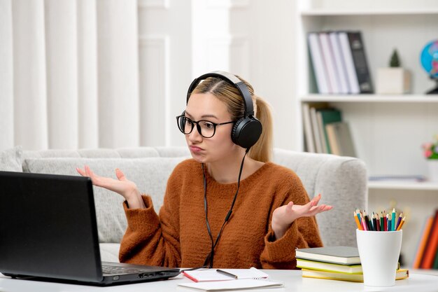 Étudiant en ligne jeune fille mignonne dans des verres et un pull orange étudiant sur ordinateur en agitant les mains