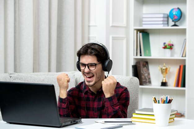 Étudiant en ligne jeune homme en chemise à carreaux avec des lunettes étudiant sur ordinateur excité avec les poings levés