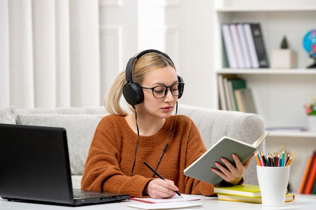 Étudiant en ligne jolie fille à lunettes et pull étudiant sur ordinateur en lisant un livre