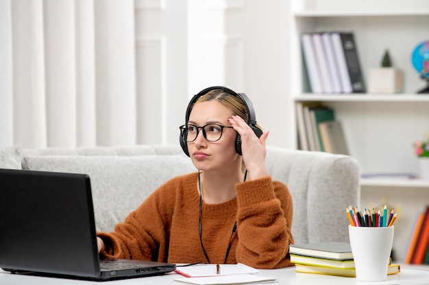 Étudiant en ligne jolie fille à lunettes et pull étudiant sur ordinateur pensant dans les écouteurs