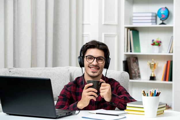 Étudiant en ligne mec mignon en chemise à carreaux avec des lunettes étudiant sur ordinateur souriant et heureux