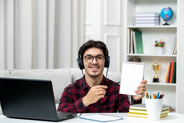 Étudiant en ligne mec mignon en chemise à carreaux avec des lunettes étudiant sur ordinateur souriant tenant des notes