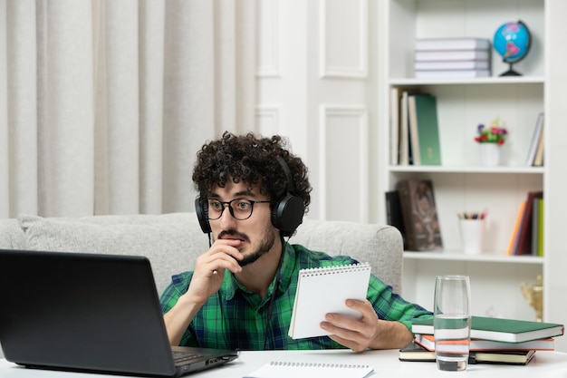 Étudiant en ligne mignon jeune homme étudiant sur ordinateur dans des verres en chemise verte se mordant le doigt