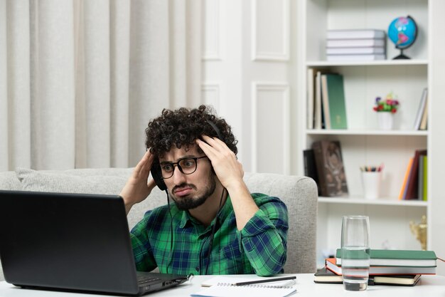 Étudiant en ligne mignon jeune homme étudiant sur ordinateur dans des verres en chemise verte tenant la tête