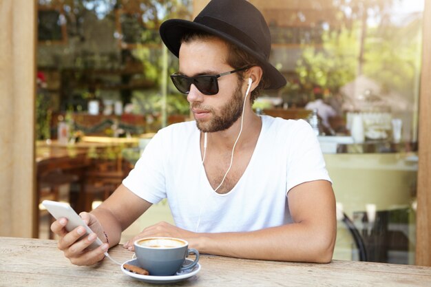 Étudiant à la mode portant des lunettes de soleil et un chapeau noir écoutant des morceaux préférés sur des écouteurs, en utilisant l'application de musique en ligne sur son téléphone mobile
