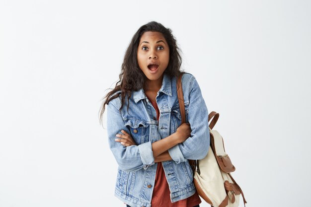 Étudiante afro-américaine surprise portant une veste en jean et un t-shirt rouge avec un sac à dos en état de choc, la bouche grande ouverte et les mâchoires tombées, gardant les bras croisés, étonnée