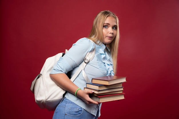 Étudiante blonde tenant une lourde pile de livres et a l'air fatiguée.