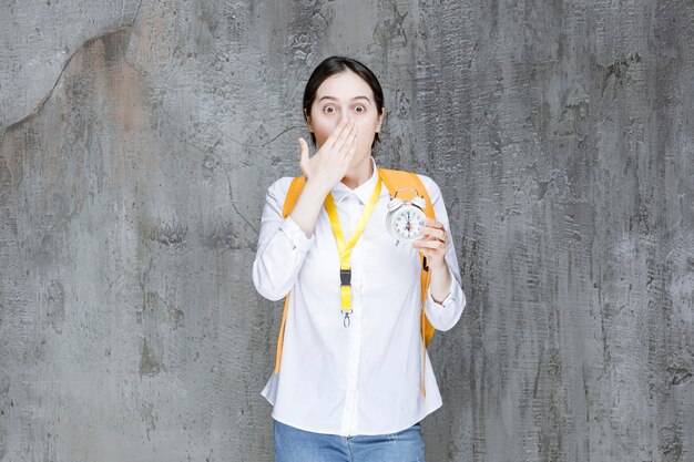 Étudiante avec sac à dos jaune tenant l'horloge et couvrant sa bouche. Photo de haute qualité