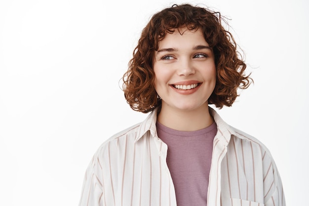 Étudiante souriante avec une coiffure courte bouclée regardant de côté la bannière du logo, se sentant heureuse et détendue, regardant à gauche l'espace de copie, debout sur fond blanc.