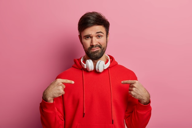 Un type caucasien mal rasé, non impressionné, se montre, demande qui moi, a une expression de visage calme, porte un sweat-shirt rouge, écoute l'audio via un casque, montre une nouvelle tenue achetée, pose sur un mur rose