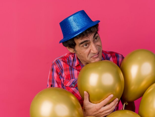 Unpleased caucasian party man wearing party hat debout derrière des ballons en saisissant un à côté isolé sur fond cramoisi