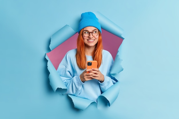 Photo gratuite l'utilisateur de la technologie d'une adolescente rousse positive tient un téléphone portable envoie des messages texte regarde de côté porte joyeusement un chapeau bleu et le cavalier regarde pensivement de côté perce le trou de papier bleu