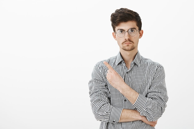 Va-t'en, ne veux pas te voir. Colère marre d'un collègue masculin européen à lunettes et chemise rayée, pointant vers l'arrière ou dans le coin supérieur gauche, mécontent et en colère, demandant à partir sur un mur gris